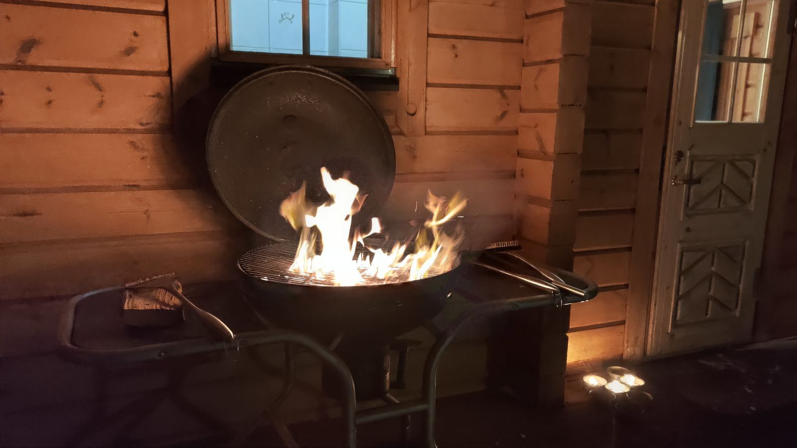 An open charcoal grill with flames leaping up, inside a wooden cabin at night.