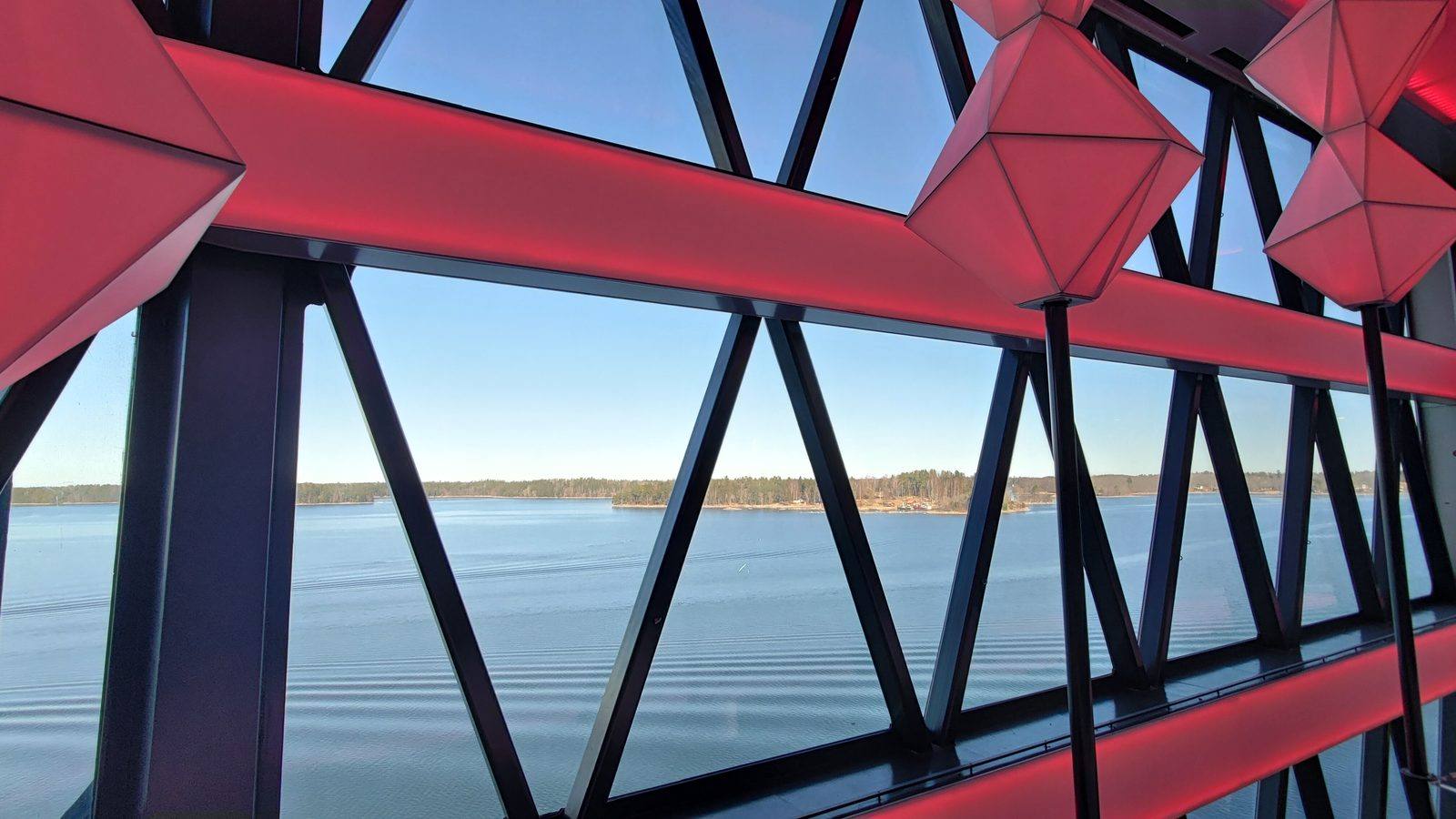 Looking out through a red-lit triangular lattice structure over a calm bay toward a forested shoreline.
